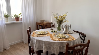 Mesa redonda con mantel a rayas, dos platos de comida, cubiertos, tetera plateada, jarrón con flores, vela. Dos sillas, ventana con plantas, mueble con libros.