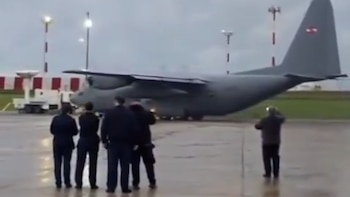 Un avión militar gris tipo Lockheed C-130 en una pista de aeropuerto mojada, con varias personas de espaldas y torres de luz al fondo
