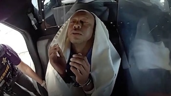 Primer plano de Tiger Woods, calvo, con ojos semicerrados y boca abierta, envuelto en una manta blanca dentro de un vehículo, junto a una persona con uniforme de sheriff