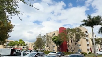 Un complejo de edificios de apartamentos color crema y rojo con un estacionamiento lleno de coches y árboles. Cielo azul con nubes blancas visibles