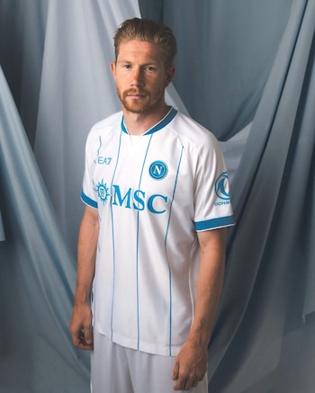 Hombre pelirrojo con barba posa. Viste camiseta de fútbol blanca con rayas y detalles azules, logotipos de marcas. Pantalones cortos blancos. Fondo de tela azul