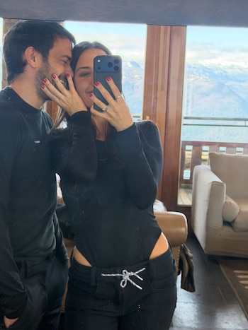 Una mujer y un hombre vestidos de negro se toman una foto en un espejo dentro de una habitación con ventanal hacia las montañas