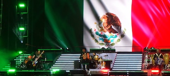 Músicos interpretan sus instrumentos en un escenario con pantallas luminosas y una imagen gigante de la bandera de México