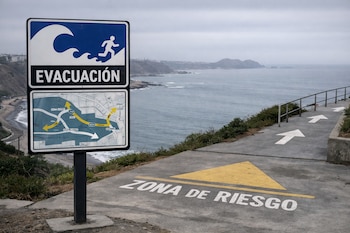 Un cartel azul y blanco de evacuación por tsunami con mapa de rutas. En el suelo, la inscripción "ZONA DE RIESGO" y flechas blancas. Mar y costa al fondo.