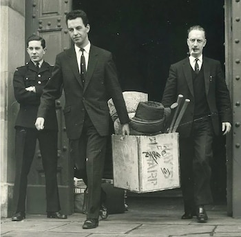 Fotografía en blanco y negro de tres hombres saliendo de una puerta. Uno es un policía uniformado, y dos hombres de traje transportan una caja de madera y otros objetos