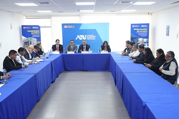 Un grupo de aproximadamente 15 personas está sentado alrededor de una mesa en forma de U cubierta con un mantel azul, en lo que parece ser una sala de reuniones