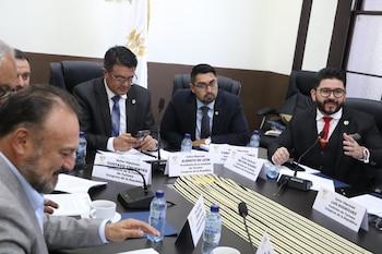 Diputados vestidos de traje participan en una mesa de trabajo con rótulos de identificación y micrófonos