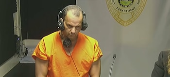Un hombre con uniforme naranja de prisión y audífonos permanece de pie frente a una pared con un escudo oficial