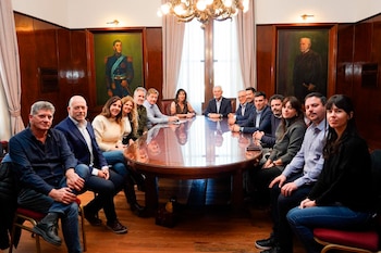 Grupo de personas sentadas alrededor de una mesa ovalada de madera en una sala con paneles de madera, dos cuadros y un candelabro