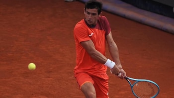 Mariano Navone, con camiseta y pantalón corto naranja y granate, golpea una pelota de tenis amarilla con una raqueta azul en una cancha de arcilla