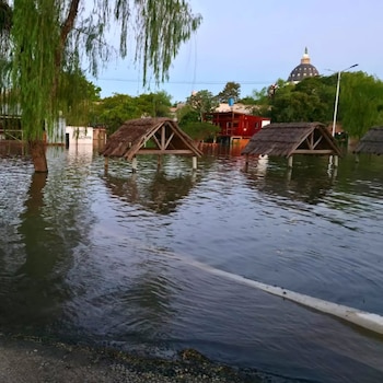 Estructuras de madera con techos de paja y árboles parcialmente sumergidos en agua turbia ante edificios y una cúpula
