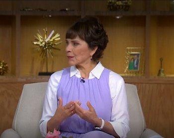 Una mujer de cabello corto, sentada en un sillón claro, viste camisa blanca y top lila, con un micrófono. Sus manos están unidas. Fondo con estanterías y adornos