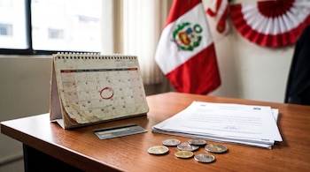 Calendario de escritorio, fecha marcada en rojo, monedas peruanas, tarjeta de pago, documentos, bandera de Perú y decoración de Fiestas Patrias.