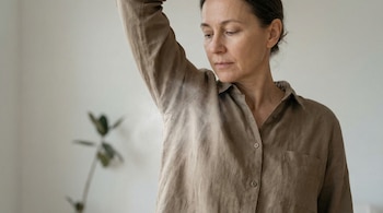 Primer plano de una mujer adulta con camisa de lino marrón, levantando el brazo derecho para revelar la axila, con una leve neblina visible. Mira hacia abajo con expresión seria.