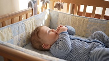 Un bebé acostado en una cuna de madera con un pijama de rayas azules y blancas se chupa el dedo pulgar. Un móvil con elefantes cuelga sobre la cuna.