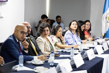 Un grupo de personas sentadas en una mesa larga con manteles azul y blanco, botellas de agua, tazas, cuadernos. Bandera de Guatemala al fondo