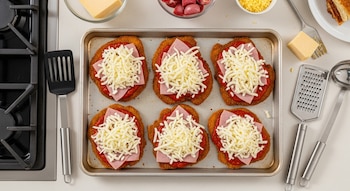 Seis milanesas napolitanas en una bandeja para horno con salsa de tomate, jamón y mozzarella rallada. Alrededor, una hornalla, espátula, rallador y cuchara.