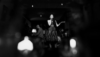 Una mujer canta en un micrófono en un escenario oscuro, con un vestido estampado y guantes. Detrás, un pianista y un baterista son visibles