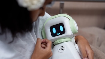 Primer plano de una niña con cabello oscuro y gorro blanco interactuando con un robot blanco y verde claro, que tiene ojos y sonrisa azules en su pantalla