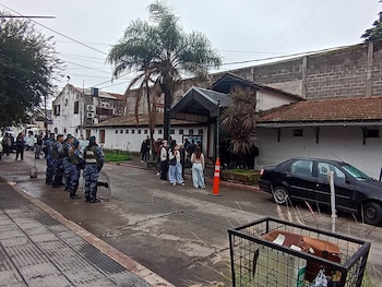 Vista exterior de un edificio con techo de tejas y un cartel de entrada oscuro. Agentes de policía y civiles de pie en una calle mojada con coches aparcados