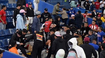 Multitud de espectadores en las gradas de asientos azules de un estadio forcejeando cerca de un agente policial.