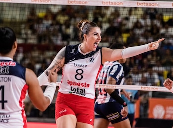 Jugadora de voleibol con uniforme blanco y rojo celebrando en la cancha, gritando y señalando, con la red de voleibol y otros jugadores detrás