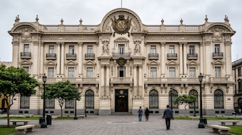 Fachada de tres pisos con columnas, balcones y emblema dorado frente a una plaza con árboles, bancos de piedra y tres transeúntes.
