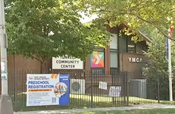 Edificio de la YMCA con letrero de 'Yurrel Community Center'. Al frente, una valla de metal y un cartel de registro para preescolar. Árboles verdes