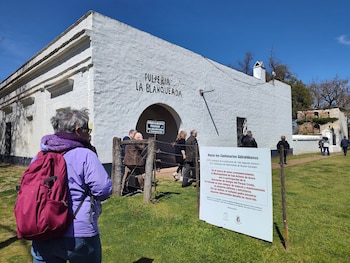 Un grupo de personas camina sobre el césped hacia un edificio blanco con el cartel Pulpería La Blanqueada bajo un cielo despejado