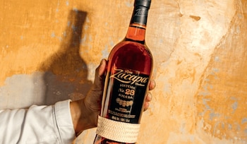 Una mano sostiene una botella de vidrio con líquido ámbar, etiqueta negra y banda de rafia, frente a una pared de fondo naranja irregular