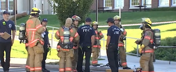 Un grupo de bomberos con trajes de protección y cascos conversa en el exterior de un edificio de ladrillos con césped al fondo
