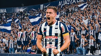 Jugador de Alianza Lima con camiseta blanquiazul celebra eufóricamente con puños cerrados y boca abierta. Detrás, hinchas vibrantes con banderas en estadio.