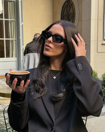 Guille Giles viste chaqueta negra y gafas de sol oscuras mientras sostiene una taza de café con leche y se toca el cabello