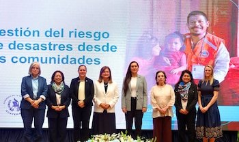 Ocho mujeres posan alineadas en un escenario frente a una pantalla azul que contiene texto e imágenes sobre gestión de riesgos