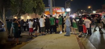 Grupo de personas en una calle concurrida de noche, frente a un local "La Poblana", con edificios iluminados y coches en la vía