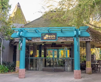 Entrada de edificio con puertas de cristal, pérgola turquesa, cartel azul "Oasis on the Rocks", techo de tejas, árboles, plantas y mobiliario exterior