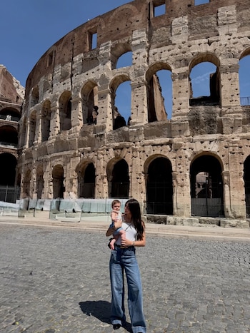 Ailén Cova, con camiseta blanca y jeans, sostiene a su hija bebé frente al Coliseo romano bajo un cielo azul claro en una calle adoquinada
