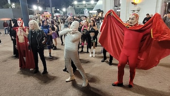 Luchadores de Titanes en el Ring, incluyendo un diablo rojo y una momia, posan entre una multitud en la Feria del Libro por la noche