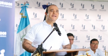 Un hombre con camisa blanca habla ante un micrófono en un atril, frente a un panel con logotipos y la bandera de Guatemala