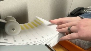 Manos organizan una pila de documentos blancos con franjas amarillas. Se ve parte de una máquina y un teléfono de oficina en segundo plano