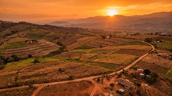 Vista aérea de terrenos agrícolas en El Salvador, con cultivos en laderas, caminos de tierra, casas, montañas en el fondo y un atardecer naranja.