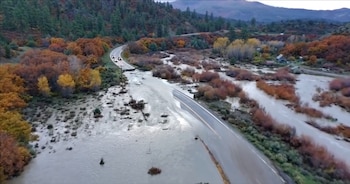 Vista aérea de una carretera inundada por agua marrón, con vehículos detenidos y colinas arboladas con follaje otoñal