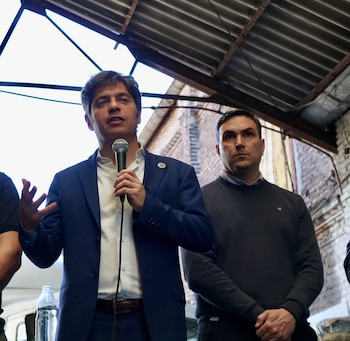 Axel Kicillof hablando en un micrófono con una mano levantada, vistiendo traje azul y camisa blanca. A su derecha, un hombre de su equipo