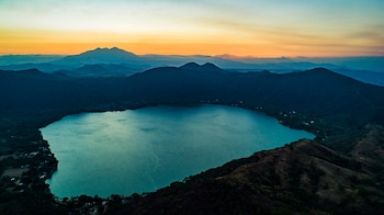 Vista aérea de una laguna azul rodeada de montañas oscuras y vegetación. El horizonte muestra tonos anaranjados y azules, con pequeñas construcciones en la orilla