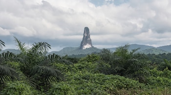 Una alta formación rocosa de cumbre estrecha sobresale entre plantas de gran tamaño y palmeras bajo un cielo nublado.