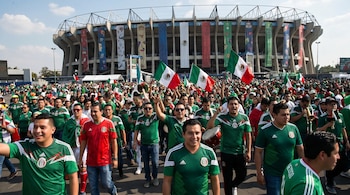 Gran multitud de aficionados con camisetas verdes y rojas de la selección mexicana, algunos agitando banderas, frente al Estadio Azteca en un día soleado.