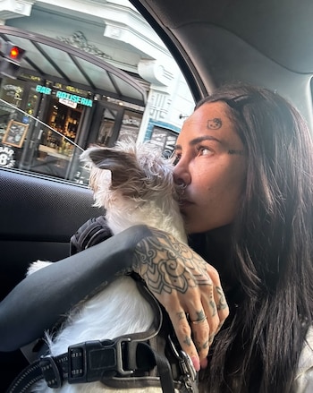 Una mujer con tatuajes en el rostro y los brazos sostiene a un perro pequeño de pelaje blanco dentro de un vehículo