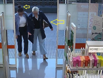 Dos mujeres con los rostros difuminados en el acceso de cristal de un establecimiento comercial con estanterías de productos y carteles visibles