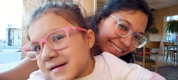 Primer plano de una niña y una mujer, ambas sonriendo y mirando a cámara. La niña usa gafas rosadas y la mujer gafas de montura clara, con un interior difuminado