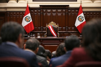 Personas sentadas desenfocadas en primer plano. Silla presidencial vacía con banda bicolor, dos banderas de Perú, mesa con micrófonos y vasos de agua, panel de madera.